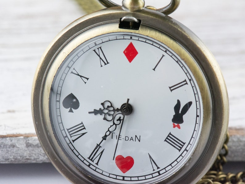 Op de afbeelding: Een vintage-stijl zakhorloge met een witte wijzerplaat en zwarte Romeinse cijfers. Het horloge heeft een vergulde kast en een ketting. De wijzerplaat van het horloge heeft speelkaartsymbolen en een konijnen-silhouet.