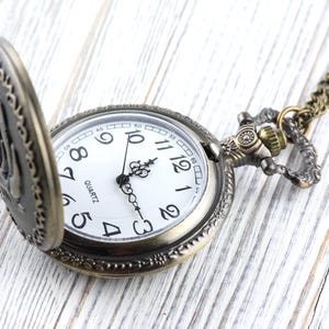 May include: Antique-style pocket watch with a bronze-colored case and chain. The watch face is white with black numerals and hands, and the word "QUARTZ" is printed at the bottom. The watch is partially open, revealing the time.