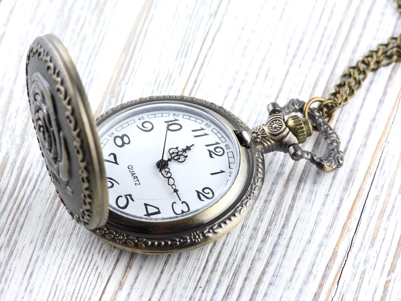 May include: Antique-style pocket watch with a bronze-colored case and chain. The watch face is white with black numerals and hands, and the word "QUARTZ" is printed at the bottom. The watch is partially open, revealing the time.