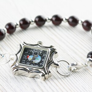 May include: A silver watch with a snowman design on the face, attached to a bracelet made of dark red beads.
