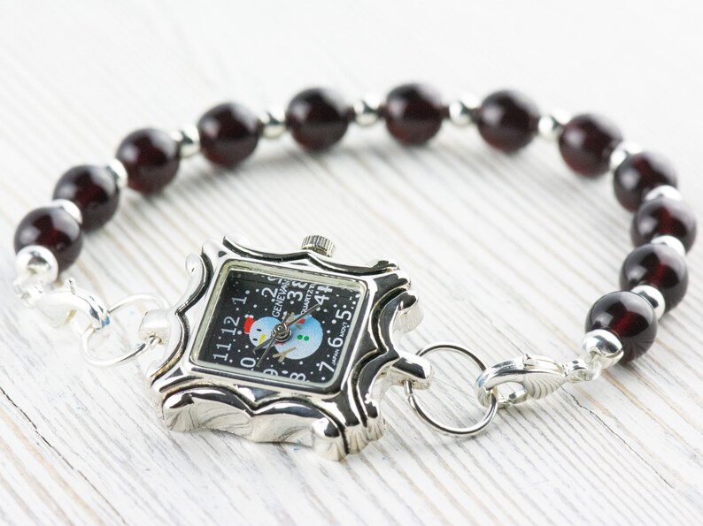 May include: A silver watch with a snowman design on the face, attached to a bracelet made of dark red beads.