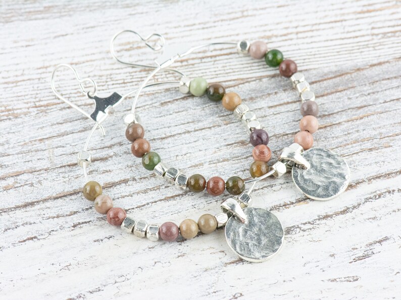 May include: A pair of silver hoop earrings with a hammered silver disc charm. The hoops are adorned with multi-colored gemstone beads.