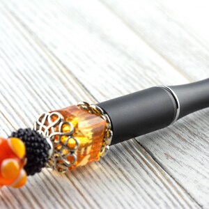 May include: A black and silver vape pen with decorative orange and silver beads. The pen has a decorative amber-colored bead with a silver filigree design. The pen is on a white wooden surface.