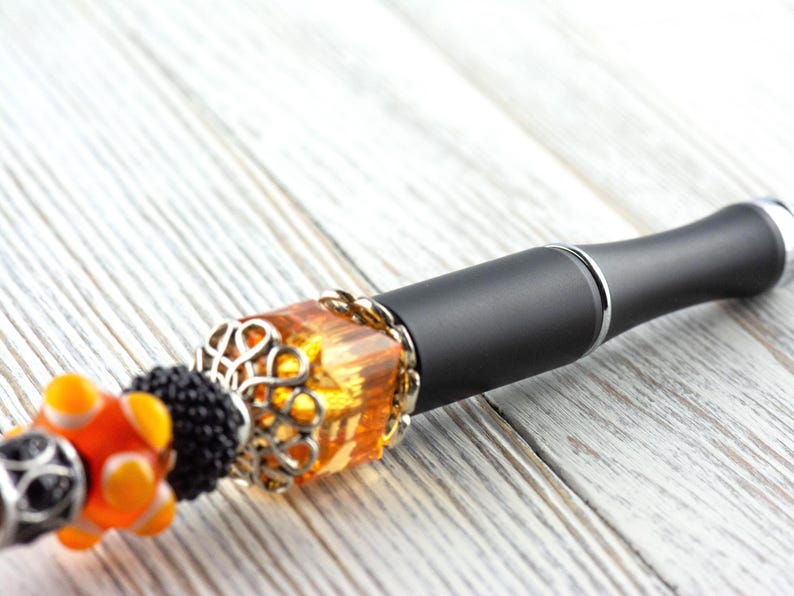 May include: A black and silver vape pen with decorative orange and silver beads. The pen has a decorative amber-colored bead with a silver filigree design. The pen is on a white wooden surface.