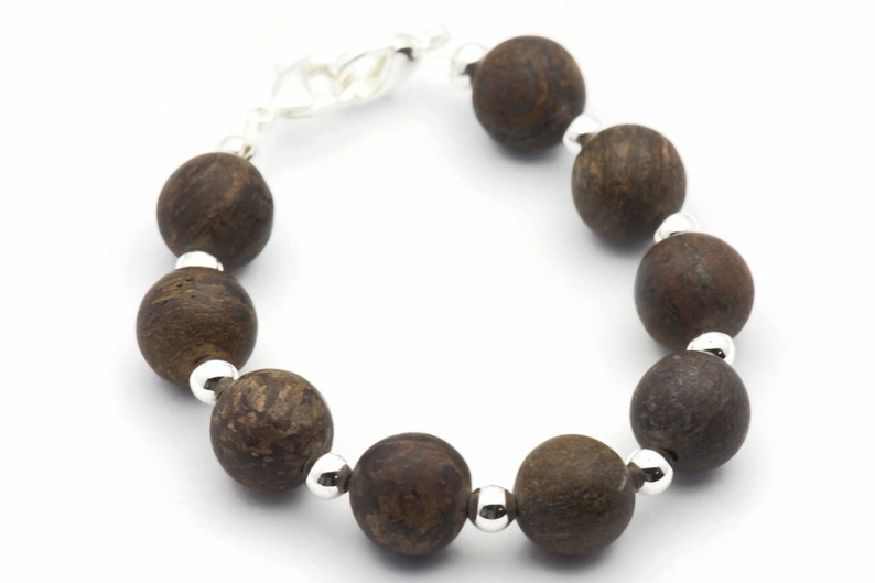 Puede incluir: Un brazalete de cuentas de madera marr&oacute;n con detalles plateados. El brazalete tiene un cierre de broche.