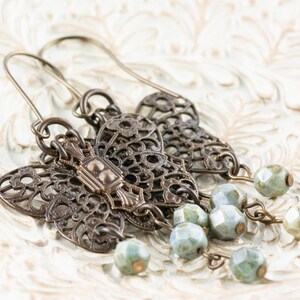May include: Antique bronze-toned earrings with intricate filigree designs. Each earring features a decorative center piece and dangling strands of faceted, pale green beads. The earrings are displayed on a textured, off-white surface.