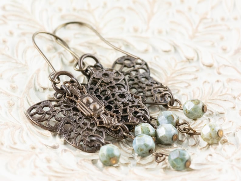 May include: Antique bronze-toned earrings with intricate filigree designs. Each earring features a decorative center piece and dangling strands of faceted, pale green beads. The earrings are displayed on a textured, off-white surface.