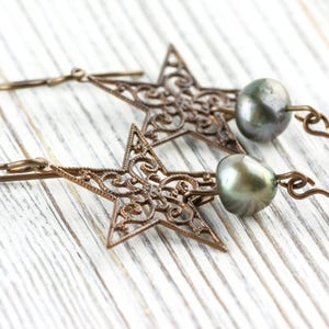 May include: A pair of bronze-colored star-shaped earrings with intricate filigree detailing. Each earring features a dark gray pearl and a clear crystal bead. The earrings are displayed on a white wooden surface.