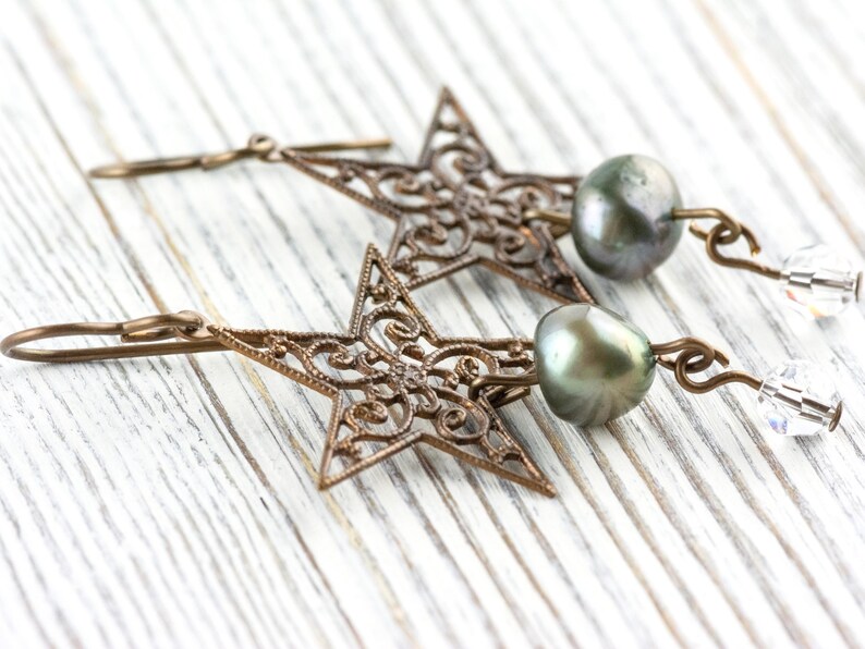 May include: A pair of bronze-colored star-shaped earrings with intricate filigree detailing. Each earring features a dark gray pearl and a clear crystal bead. The earrings are displayed on a white wooden surface.