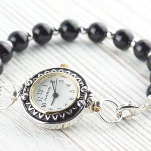 Pu&ograve; includere: Un orologio argentato e dorato con quadrante bianco e numeri neri, collegato a un braccialetto di perline nere. Il quadrante dell'orologio ha il marchio "NARMI". Il braccialetto &egrave; assicurato con una chiusura argentata.