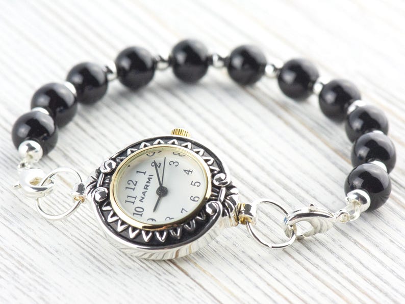 Pu&ograve; includere: Un orologio argentato e dorato con quadrante bianco e numeri neri, collegato a un braccialetto di perline nere. Il quadrante dell'orologio ha il marchio "NARMI". Il braccialetto &egrave; assicurato con una chiusura argentata.