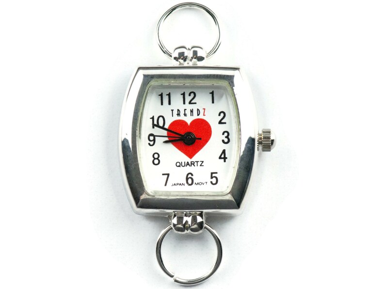 Puede incluir: Reloj de cuarzo plateado con esfera blanca con un coraz&oacute;n rojo y n&uacute;meros negros. El reloj tiene forma rectangular con bordes redondeados y dos llaveros. El reloj dice "TRENDZ" y "QUARTZ".