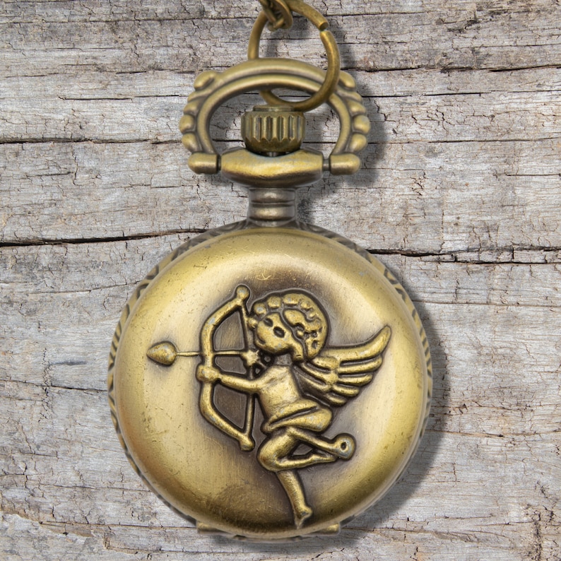 Puede incluir: Un reloj de bolsillo dorado con una cadena. La esfera del reloj presenta un grabado detallado de un querub&iacute;n sosteniendo un arco y una flecha.
