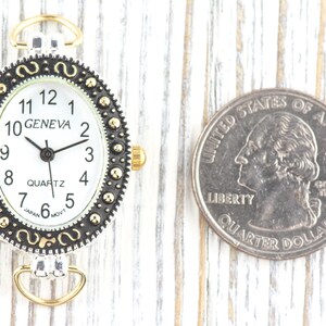 May include: Oval Geneva watch face with a white dial, black hands, and gold-tone accents. The watch face is next to a U.S. quarter for size comparison. The watch has a silver-tone frame with a decorative border.