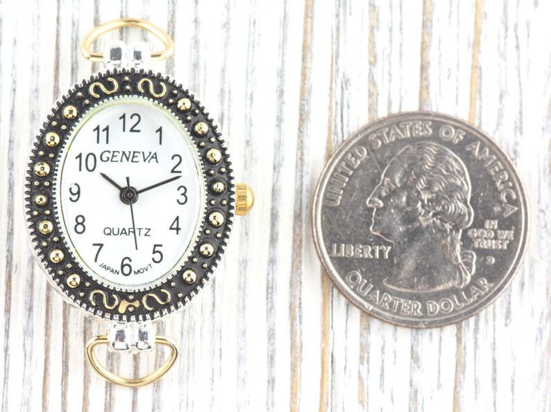 May include: Oval Geneva watch face with a white dial, black hands, and gold-tone accents. The watch face is next to a U.S. quarter for size comparison. The watch has a silver-tone frame with a decorative border.