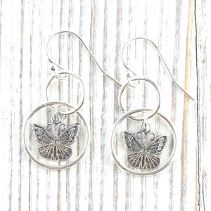 May include: Silver-toned butterfly earrings. Each earring features a circular frame with a butterfly pendant. The butterfly has detailed wing patterns and a delicate design. The earrings hang from a simple hook.