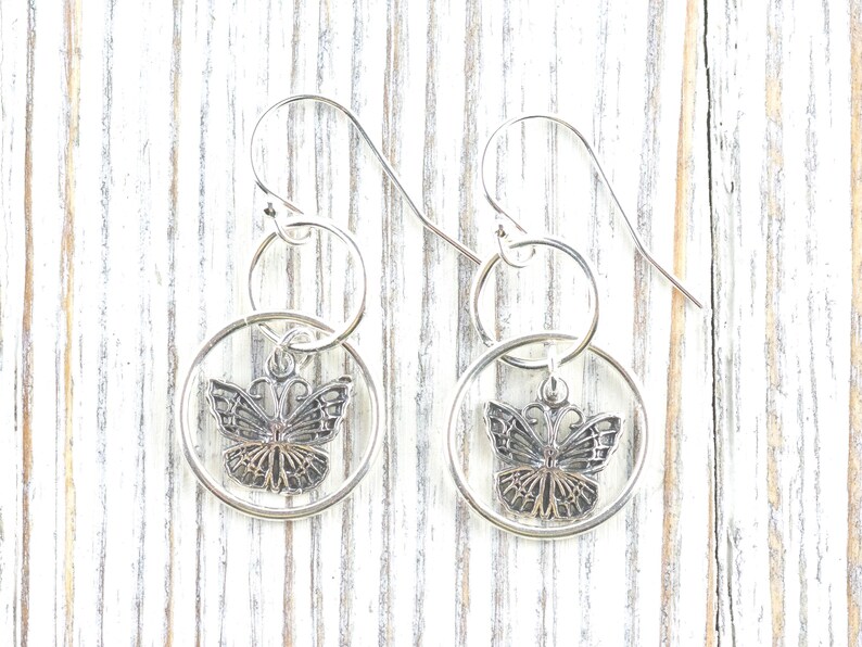 May include: Silver-toned butterfly earrings. Each earring features a circular frame with a butterfly pendant. The butterfly has detailed wing patterns and a delicate design. The earrings hang from a simple hook.