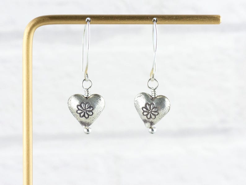 K&ouml;nnte beinhalten: Ein Paar silberfarbene herzf&ouml;rmige Ohrringe. Jeder Ohrring hat einen Herzanh&auml;nger mit floralem Design und einer kleinen Perle unten. Die Ohrringe h&auml;ngen an d&uuml;nnen, gebogenen Silberdr&auml;hten. Pr&auml;sentiert vor einem wei&szlig;en Hintergrund.