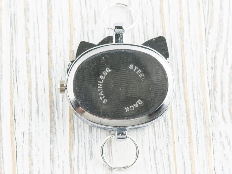 May include: The back of a silver watch with a round face and a black band. The watch has the words "Back", "Stainless Steel" engraved on the back.