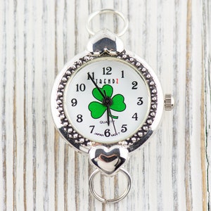 Peut inclure: Montre argent&eacute;e avec un cadran blanc et un motif de tr&egrave;fle &agrave; quatre feuilles vert. La montre a un fermoir en forme de c&oelig;ur et affiche "TREND1" sur le cadran.