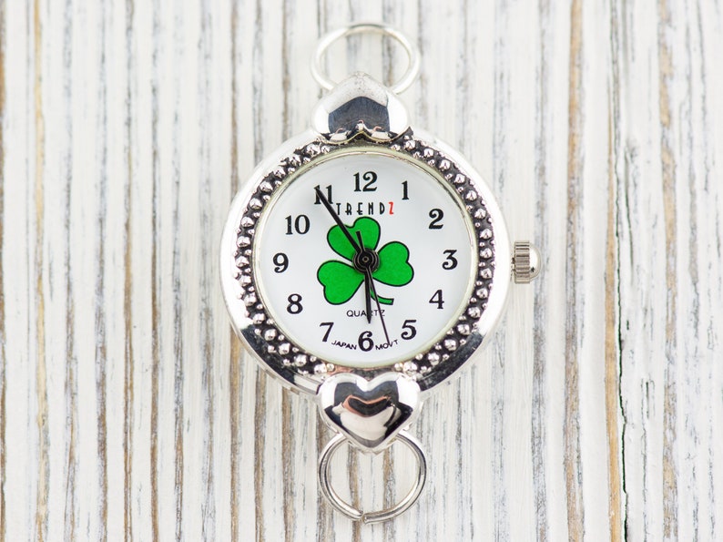 Peut inclure: Montre argent&eacute;e avec un cadran blanc et un motif de tr&egrave;fle &agrave; quatre feuilles vert. La montre a un fermoir en forme de c&oelig;ur et affiche "TREND1" sur le cadran.