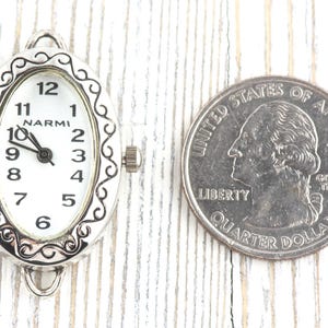 May include: An oval watch face with a white dial and black numerals, marked "NARMI." The silver-toned frame has a decorative border. A U.S. quarter is next to the watch face for size comparison.