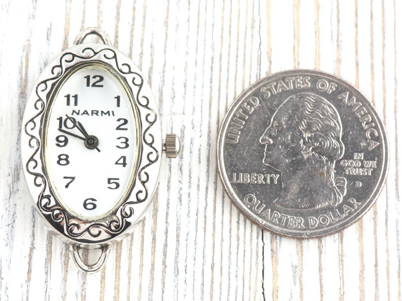 May include: An oval watch face with a white dial and black numerals, marked "NARMI." The silver-toned frame has a decorative border. A U.S. quarter is next to the watch face for size comparison.