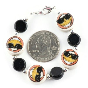 May include: A bracelet with black and white ceramic beads featuring a cartoon duck wearing sunglasses. The bracelet is silver and has a lobster clasp.