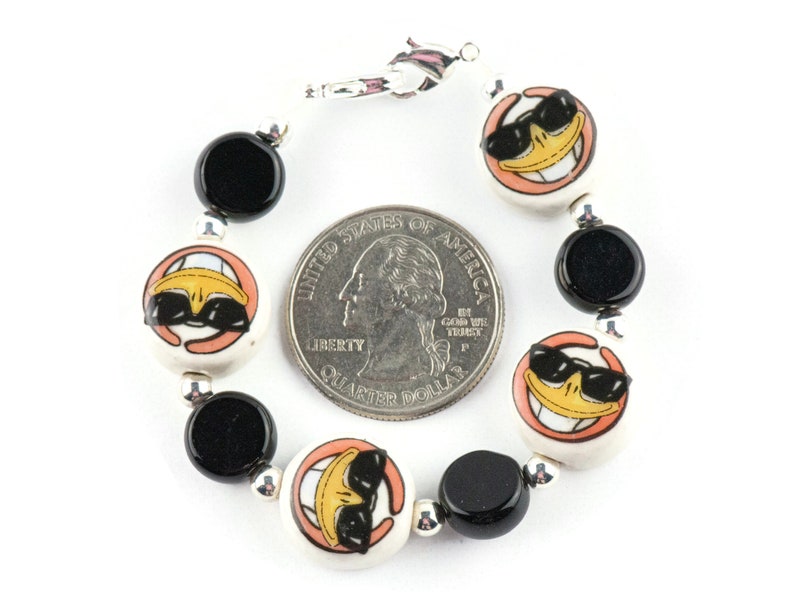 May include: A bracelet with black and white ceramic beads featuring a cartoon duck wearing sunglasses. The bracelet is silver and has a lobster clasp.