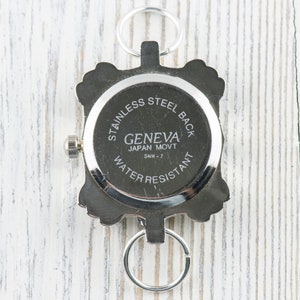 May include: The back of a silver watch with a square face. The watch face says "Geneva Japan Movt SMW-7" and "Stainless Steel Back Water Resistant".