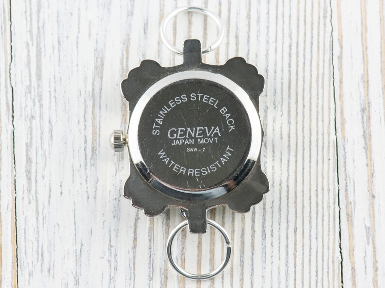 May include: The back of a silver watch with a square face. The watch face says "Geneva Japan Movt SMW-7" and "Stainless Steel Back Water Resistant".