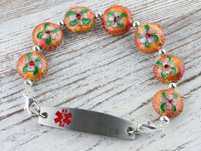 May include: A medical alert bracelet with a silver chain and orange and green floral beads. The bracelet has a silver tag that reads "Type 1 Diabetes".