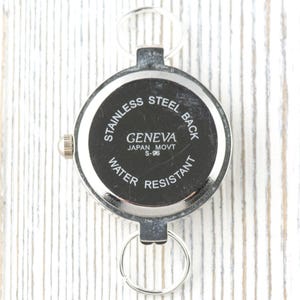 May include: A silver Geneva watch with a black face, displaying "STAINLESS STEEL BACK", "GENEVA JAPAN MOVT S-96", and "WATER RESISTANT". It features a small crown and two key rings for attachment.