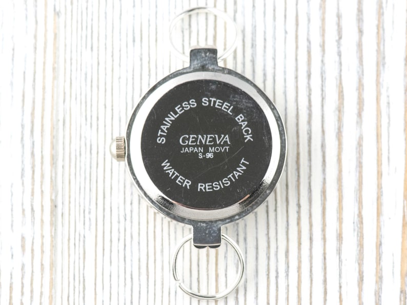 May include: A silver Geneva watch with a black face, displaying "STAINLESS STEEL BACK", "GENEVA JAPAN MOVT S-96", and "WATER RESISTANT". It features a small crown and two key rings for attachment.