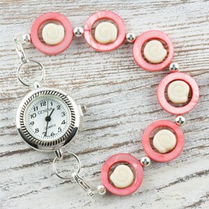 Pu&ograve; includere: Un orologio da tasca argentato con quadrante bianco e numeri neri. L'orologio &egrave; attaccato a un braccialetto di perline rosa e bianche.