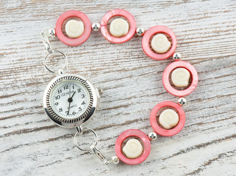 Pu&ograve; includere: Un orologio da tasca argentato con quadrante bianco e numeri neri. L'orologio &egrave; attaccato a un braccialetto di perline rosa e bianche.