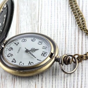 May include: Antique brass pocket watch with a white face, black numerals, and ornate hands. The watch has a hinged cover with a decorative design and a matching chain. The watch is open, revealing the time.