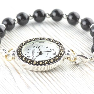 May include: A Geneva quartz watch with a white face and black hands, set in a silver-tone case with gold accents. The watch is attached to a bracelet made of black, round beads. The watch is connected with silver-tone clasps.