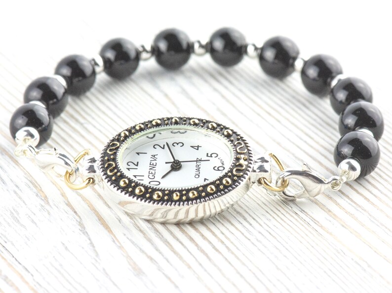 May include: A Geneva quartz watch with a white face and black hands, set in a silver-tone case with gold accents. The watch is attached to a bracelet made of black, round beads. The watch is connected with silver-tone clasps.