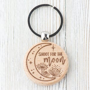 May include: Wooden keychain with a crescent moon design and the text "Shoot for the moon". The keychain has a silver metal ring.