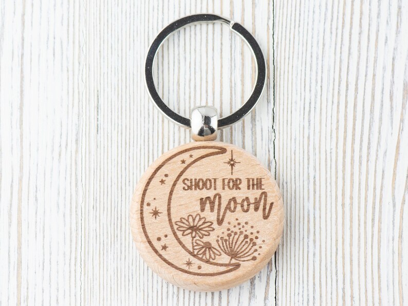 May include: Wooden keychain with a crescent moon design and the text "Shoot for the moon". The keychain has a silver metal ring.