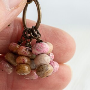 Op de afbeelding: Close-up van een handgemaakte oorbel met een cluster van kleine, onregelmatig gevormde kralen. De kralen zijn in tinten roze, bruin en beige, geregen met antiek bronzen metalen onderdelen. De oorbel wordt in een hand gehouden.