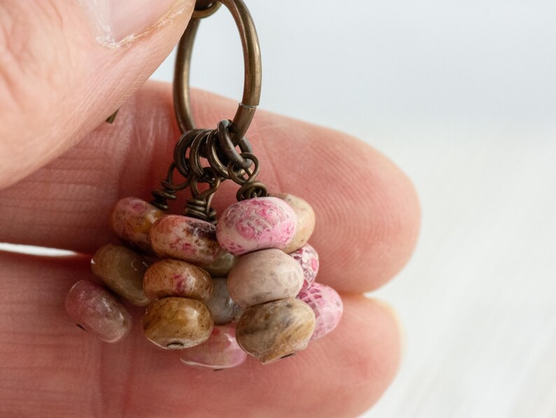 Op de afbeelding: Close-up van een handgemaakte oorbel met een cluster van kleine, onregelmatig gevormde kralen. De kralen zijn in tinten roze, bruin en beige, geregen met antiek bronzen metalen onderdelen. De oorbel wordt in een hand gehouden.