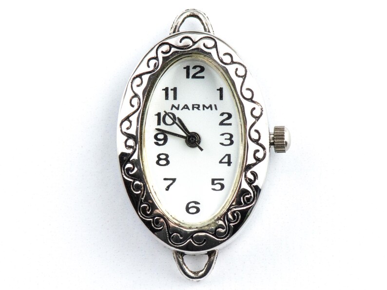 May include: Silver oval watch face with a white dial and black hands. The watch face has a decorative filigree border and the brand name "NARMI" is printed above the centre. The watch shows the time as 10:09.