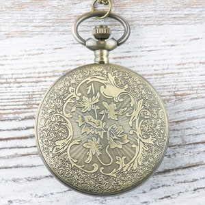 Op de afbeelding: Een gouden zakhorloge met een ingewikkeld bloemmotief aan de voorkant. Het horloge heeft een rond gezicht en een ketting die aan de bovenkant is bevestigd.