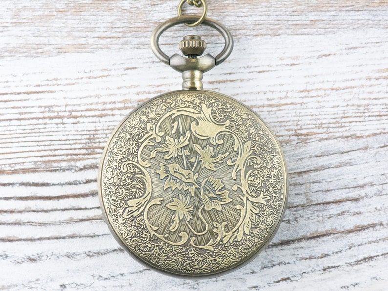 Op de afbeelding: Een gouden zakhorloge met een ingewikkeld bloemmotief aan de voorkant. Het horloge heeft een rond gezicht en een ketting die aan de bovenkant is bevestigd.