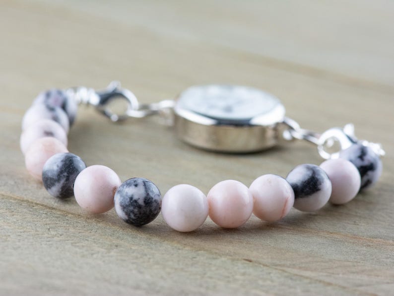May include: A silver watch with a white face attached to a bracelet made of pink and black beads.