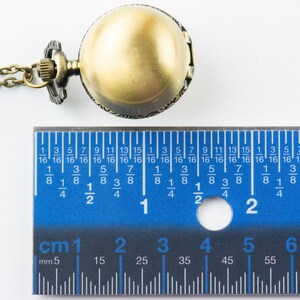 May include: A gold-toned pocket watch with a chain, measuring approximately 2 cm in diameter. The watch is on a white surface with a ruler for scale.