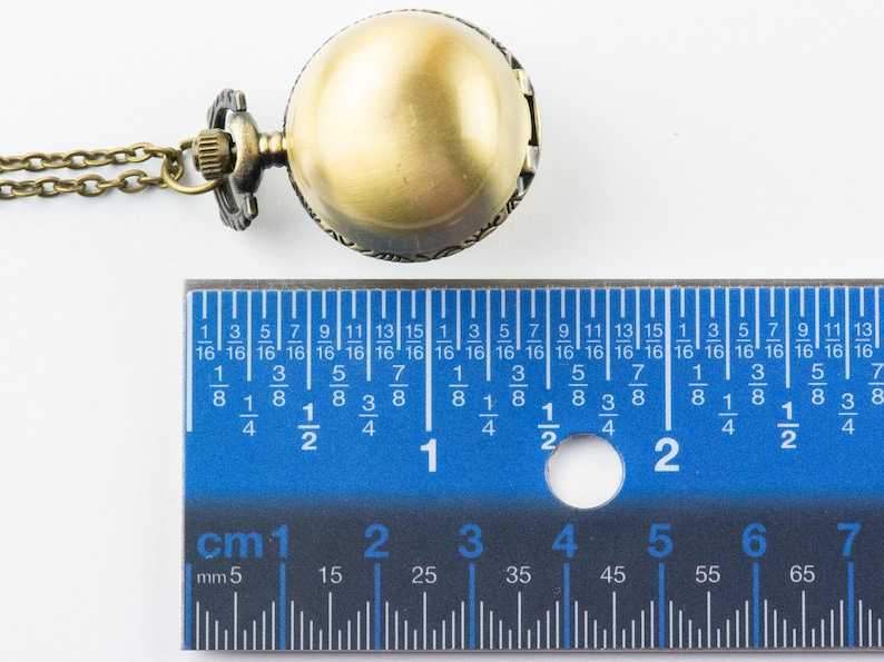 May include: A gold-toned pocket watch with a chain, measuring approximately 2 cm in diameter. The watch is on a white surface with a ruler for scale.