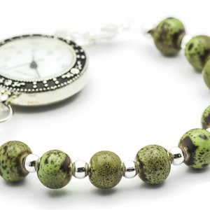 May include: A silver pocket watch with a white face and black markings attached to a green and brown beaded bracelet.